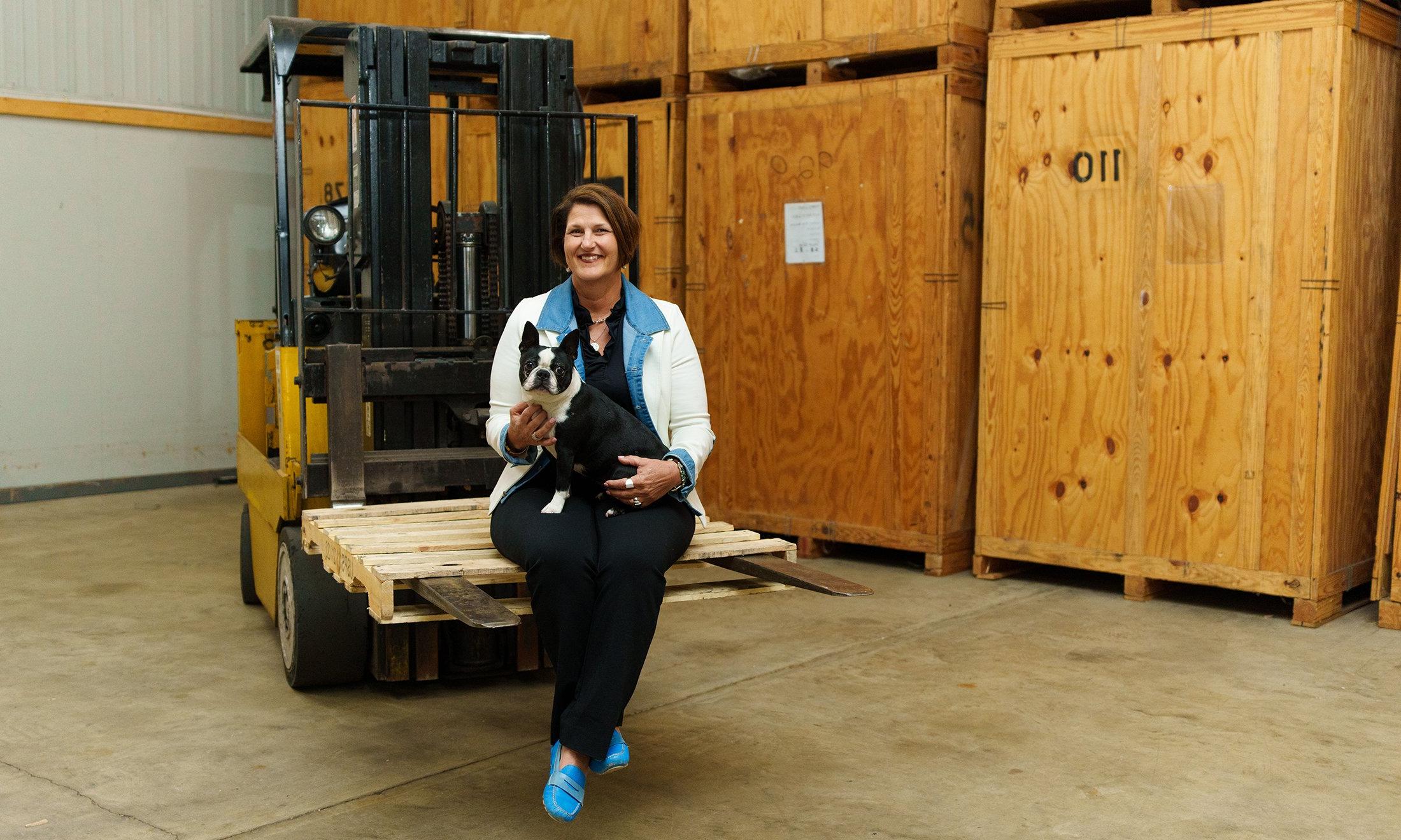 A woman holding her dog, sitting on a pallet that's on a forklift